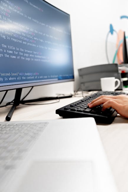 A close-up view of a silver laptop placed on a wooden table, displaying lines of code and programming text on its screen. The background is blurred but suggests a cozy indoor environment with warm lig
