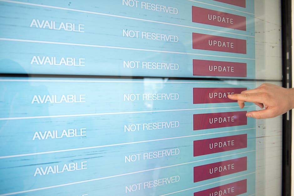 A person's hand with a neatly trimmed nail and light skin tone pointing at a digital screen displaying rows of availability statuses and red update buttons, possibly at a garden or maintenance schedul